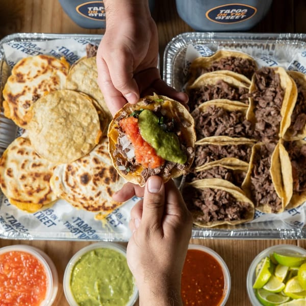 Hands holding a Tacos On The Street taco with salsa and guacamole, surrounded by more tacos and quesadillas.