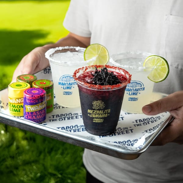 A person holding a tray with two lime margaritas, a Mezcalita Jamaica, and Twang snack salts.