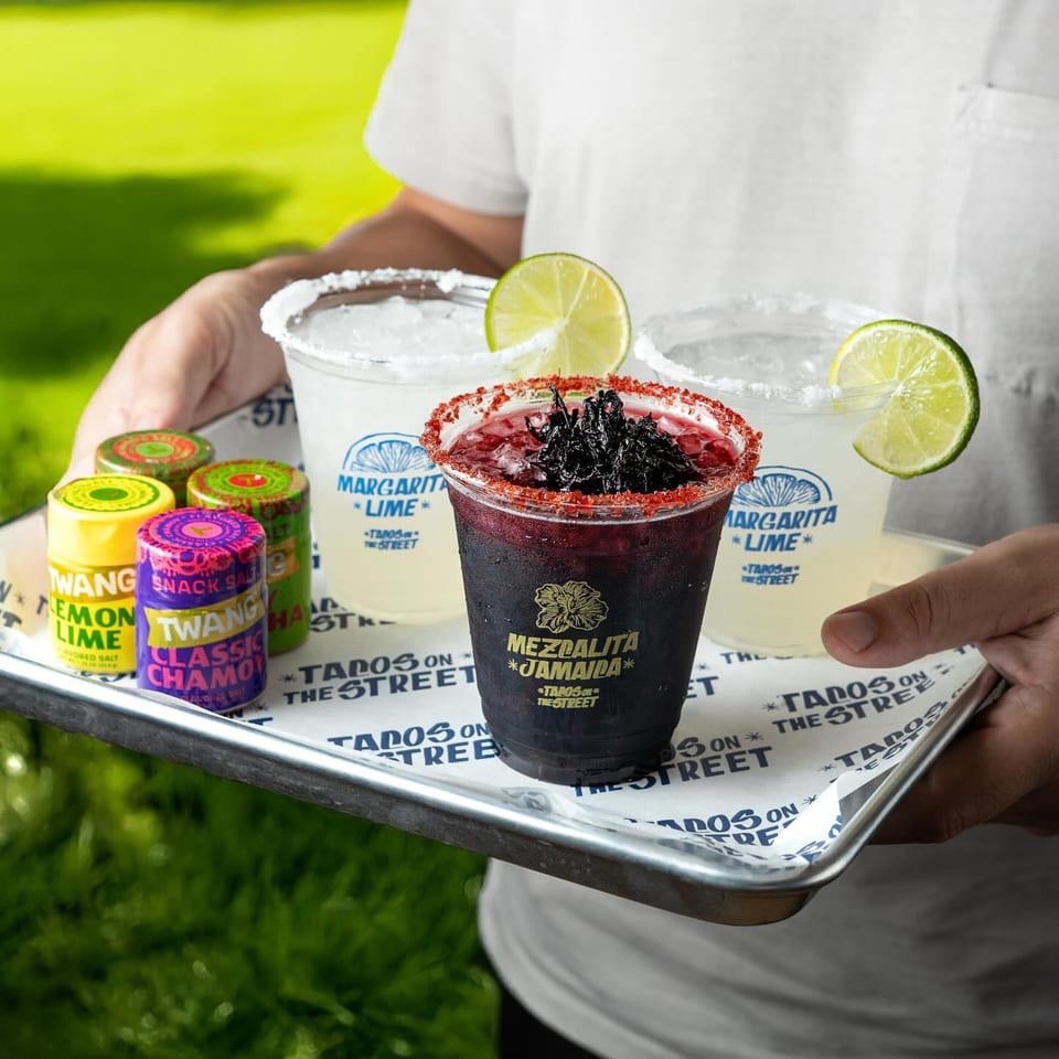 A person holding a tray with two lime margaritas, a Mezcalita Jamaica, and Twang snack salts.